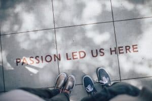 Photograph of two people standing on pavement, taken from above. Written on the ground is 'Passion Led Us Here'.