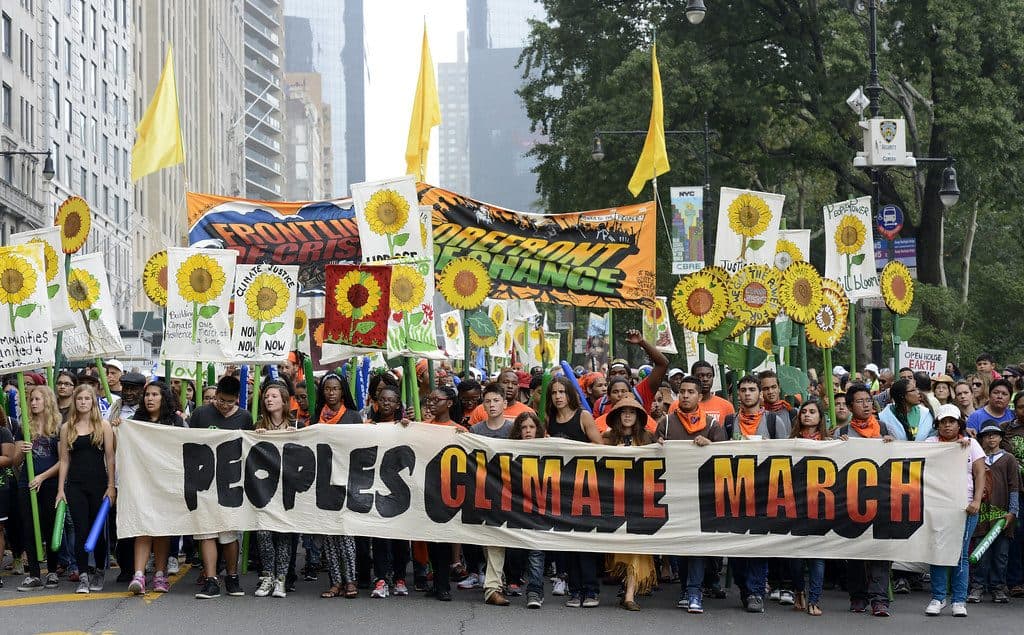 A large crowd marches behind a banner reading 'Peoples Climate March'. The march is led by First Nations people. Marchers hold colourful placards depicting sunflowers. A large banner is held high reading 'Frontlines of Impact, Forefront of Change'.