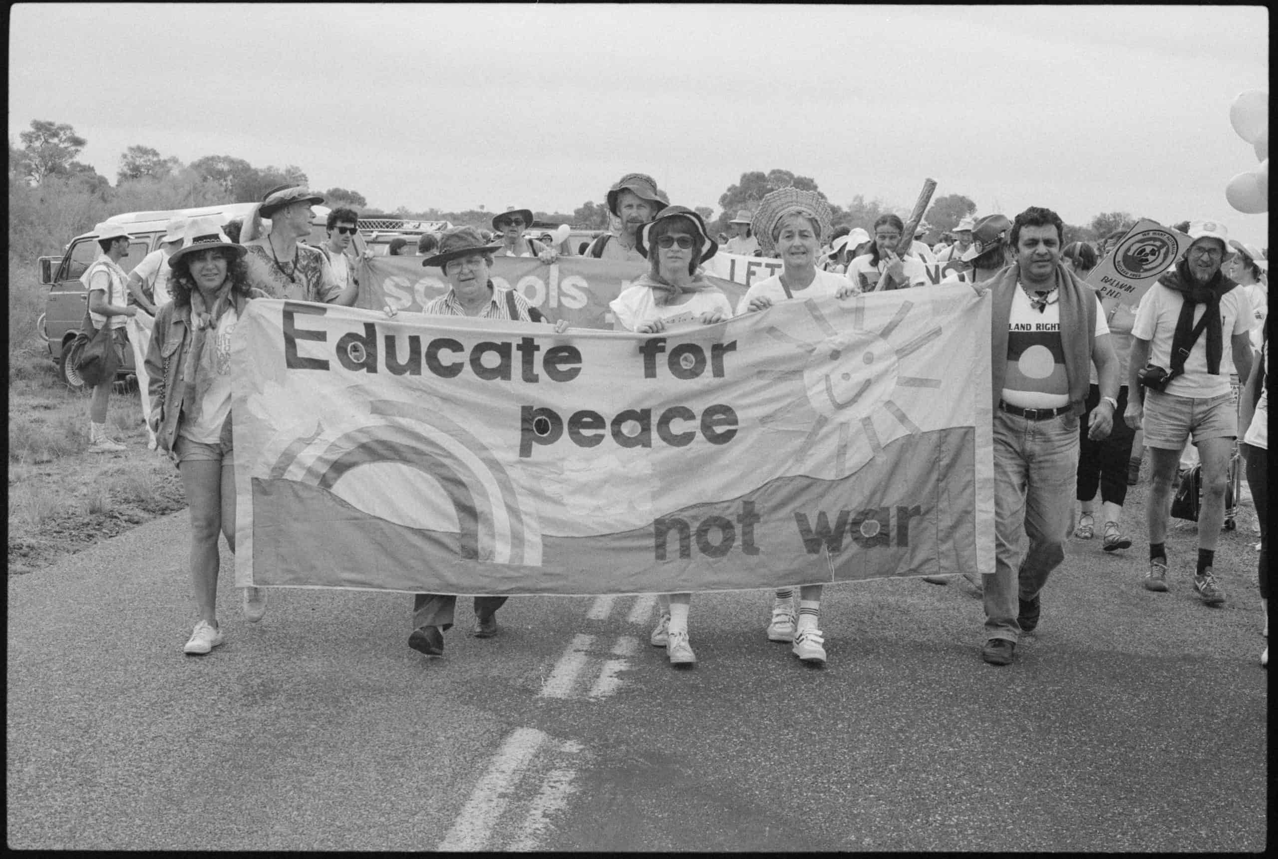 protestors walking along road carrying banner with a picture of a rainbow that reads Educate for peace not war