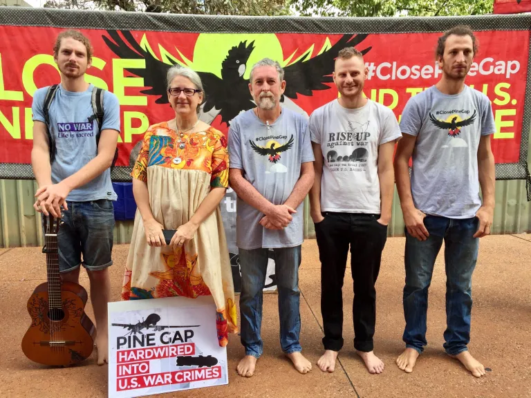A group of protestors stand in front of a banner that reads 'Close Pine Gap.' There is a black cockatoo on the banner. Another sign at the foot of the protestor reads 'Pine Gap Hard wired into US war crimes'. Four out of the five protestors are wearing anti war tshirts.