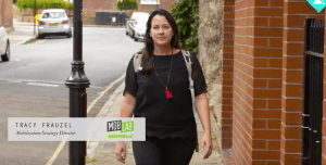 Tracey Frauzel, the Mobilisation Strategy Director who gives this course, wears black clothes with a red necklace, and walks down a street with brick walls to one side.