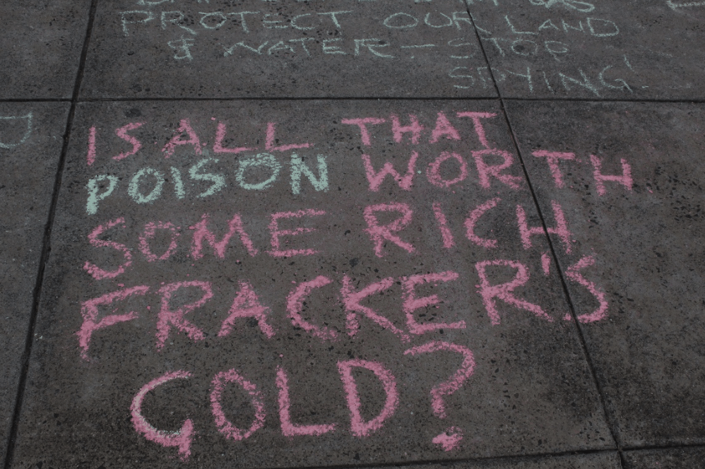 Chalk on the pavement reads 'Is all that poison worth some rich fracker's gold?'