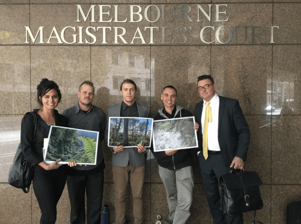 Four people stand outside the Melbourne Magistrates Court. They are smiling and holding photos of forests.