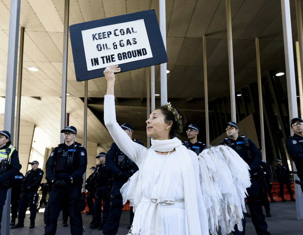 woman dressed as an angel holding up sign that says Keep oil and gas in the ground with a row of policeman standing behind her