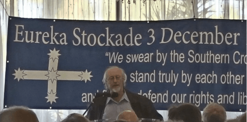 man standing in front of a eureka stockade flag giving a speech