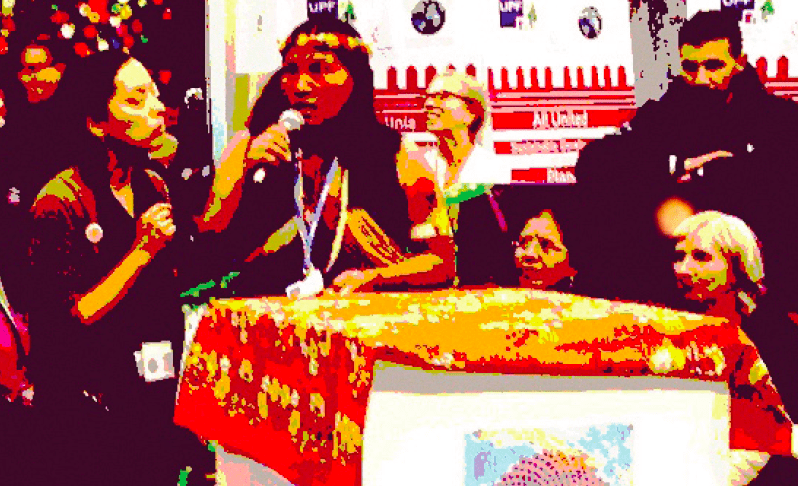 A group of people stand at the front of a forum. A speaker stands at a podium holding a microphone and wearing traditional Native American clothes. The podium has a brightly coloured fabric laid on top.