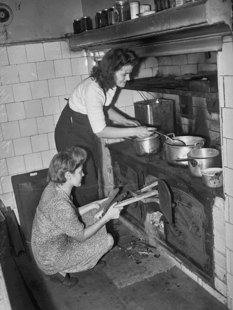 Two women lighting fire in stove in kitchen and cooking with pots and pans.