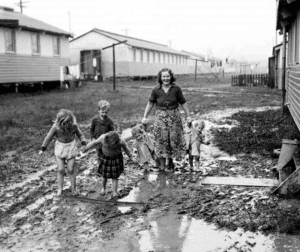 Smiling woman and 5 children walking with no shoes through mud with housing camp in the background.