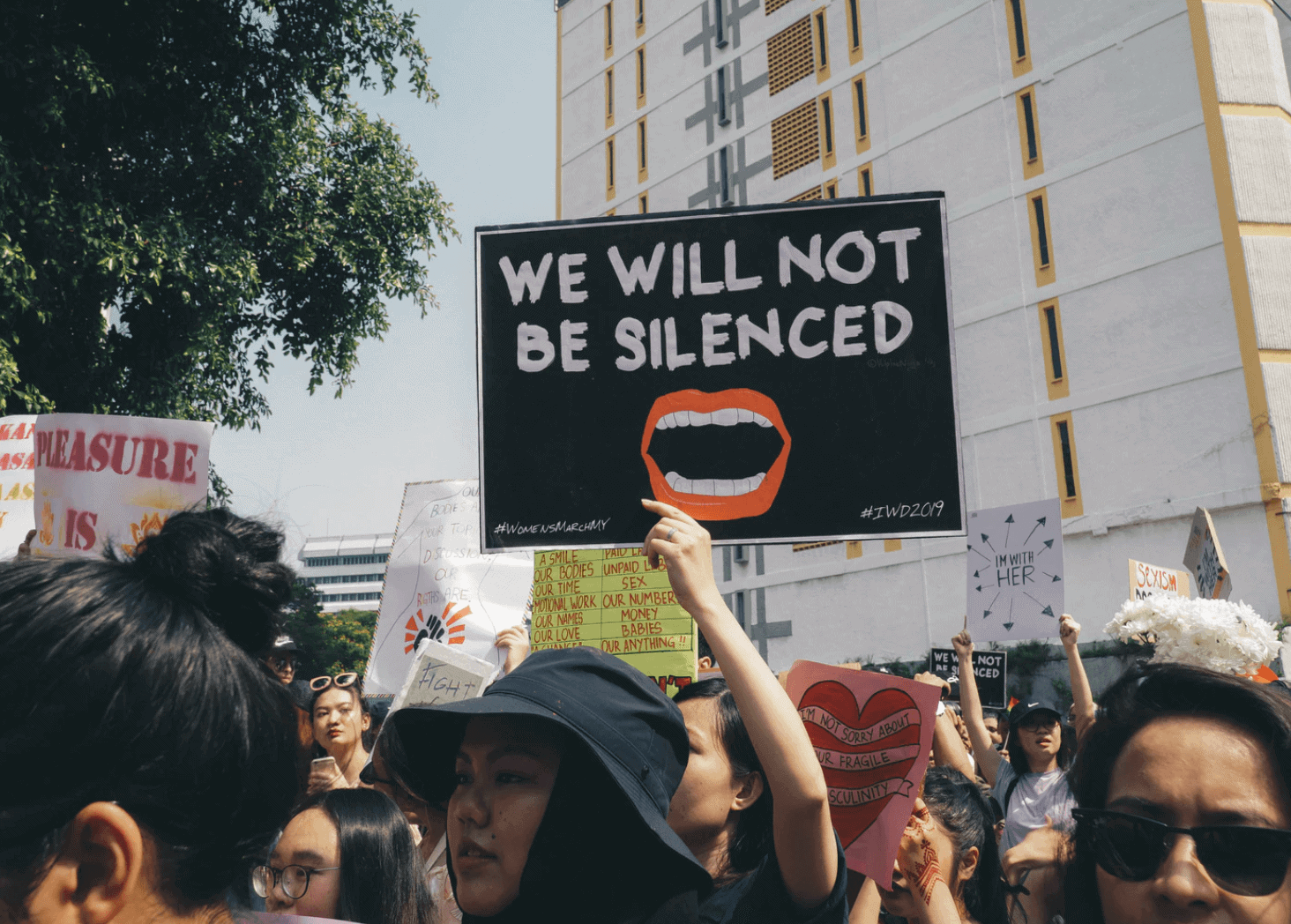 Woman holding sign up that says We will not be silenced with a picture of an open mouth. She is surrounded by other grassroots activists and protestors.