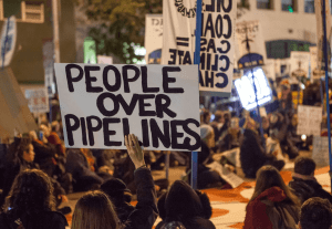 Protesters against the Dakota Access Pipeline and Keystone XL Pipeline hold a sit-in in the street next to the San Francisco Federal Building. The sign at the front says 'People over pipelines'.