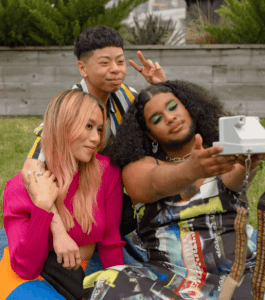 Three friends of varying genders taking a selfie in a park with a polaroid camera