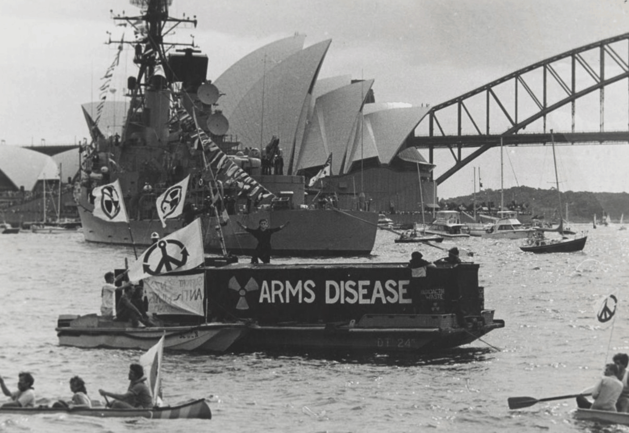 A protest boat in front of the Sydney Opera House and Harbour Bridge with a peace symbol flag and on the side of the boat it is written - Arms Disease.