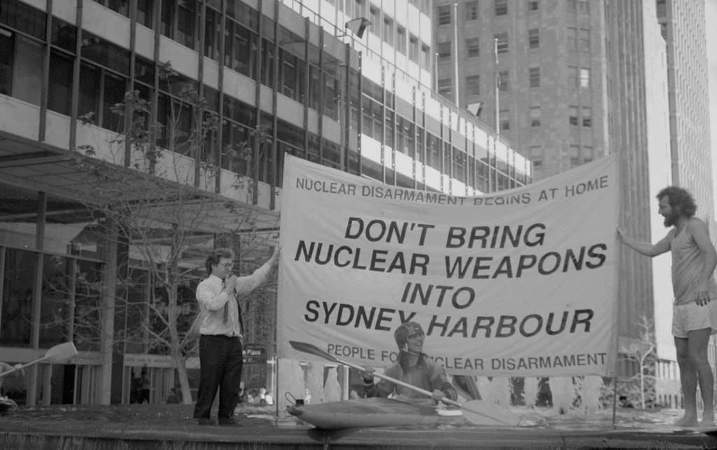 Protestors in Sydney street holding banner that says - Don't bring Nuclear Weapons into Sydney Harbour. In front of the banner is a person sitting in a kayak on a flotilla.