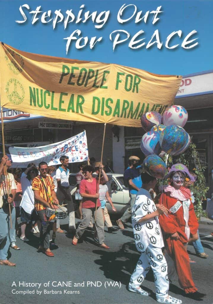 A group of people walking down the street holding banners and protesting. One big banner says Stepping Out for Peace, People for Nuclear Disarmament. Two protestors at the front are dressed in clown costumes.