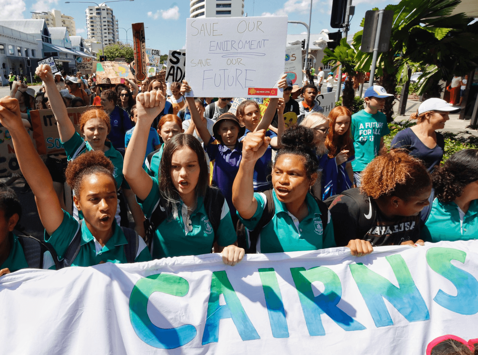 Students marching down street in protest with banner that reads Cairns. Many of them are holding their hand up in the air in a fist shape.