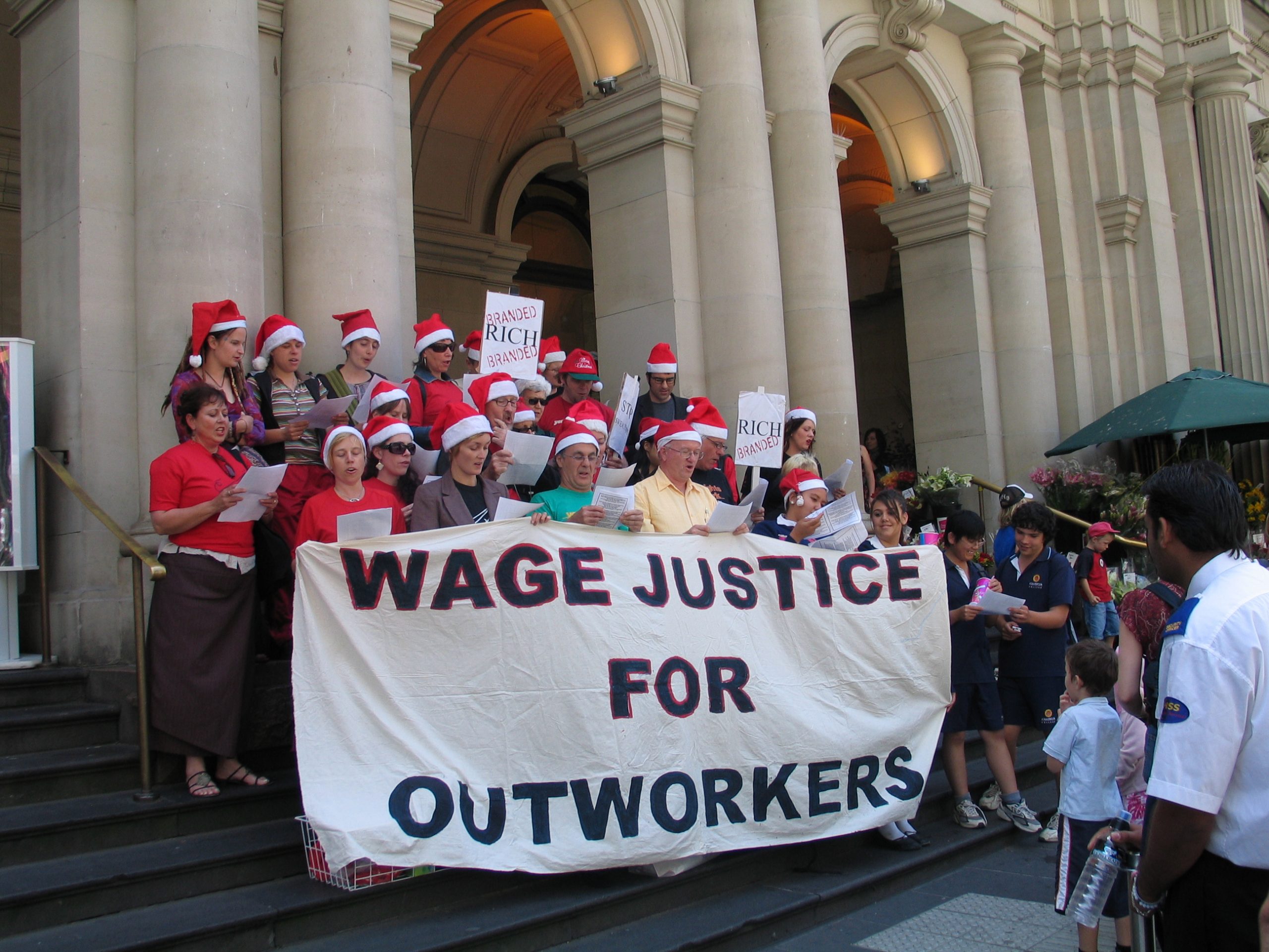 Protestors singing carols outside on steps. They are wearing santa hats and holding a banner that says Wage Justice for Outworkers.
