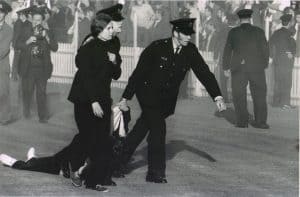 black and white photo of two police dragging one person along the ground and accompanying another woman off a sports oval