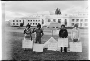Black and white photograph of four protestors standing in front of Old Parliament House. Placards read 'Land Ownership Not Lease', 'Land Rights or Else!', 'Why Pay to Use Our Own Land', 'Which Do You Choose: Land Rights or Bloodshed?'. 'Legally This is Our Land. We Shall Take It If Need Be'.