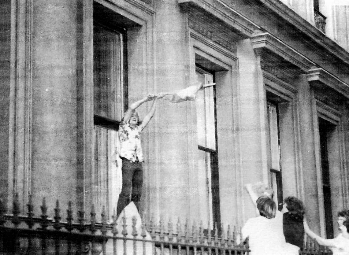 Man standing on top of iron fence waving flag outside a building.|