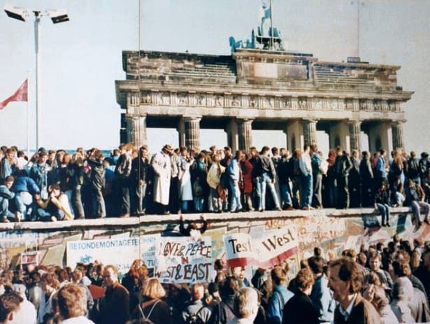 A large crowd of people standing in front of and on the Berlin Wall with the Brandenberg Gate in the background.