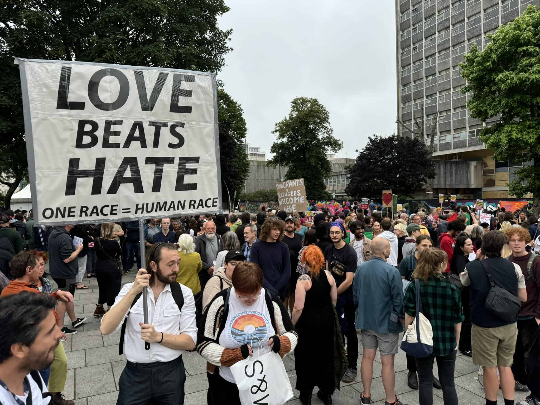 Photo of people at an anti-racist rally in the UK. A person at the front of the photo holds a large placard 'Love Beats Hate. One Race = Human Race'
