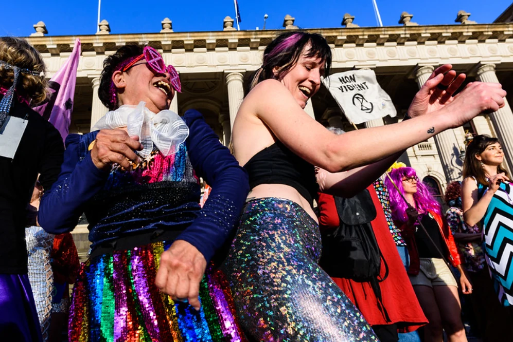 Extinction Rebellion members in disco outfits dance outside of the Victorian Parliament.|Two women wearing 1950s style clothes and pearl jewellery have faces painted black like they have used coal as make-up.||Ride Against Uranium poster.|Cyclists at a critical mass event hold their bicycles above their heads.|People dressed in costumes with a placard reading 'Libs 4 Famine