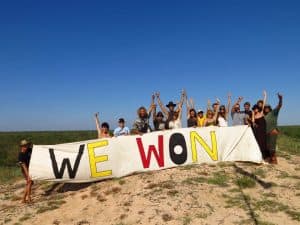 A jubilant group of people pose behind a large banner reading 'We won'. They are at the beach with sky and ocean in the background.