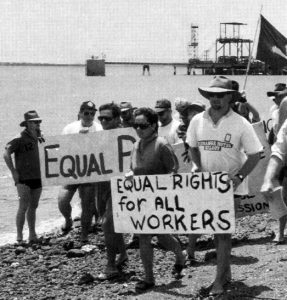 Strikers walking along a rocky beach next to the water holding signs. One reads Equal rights for all workers.