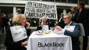 Three people at a table outside with white tablecloth that has word BlackRock on it. The people are eating fake money. A banner in the background says When the last tree is dead, the last fish is caught and the last river is poisoned, you will realise that you cannot eat money. The woman seated on the left wears a sign on her chest that reads BlackRock is the real enemy.