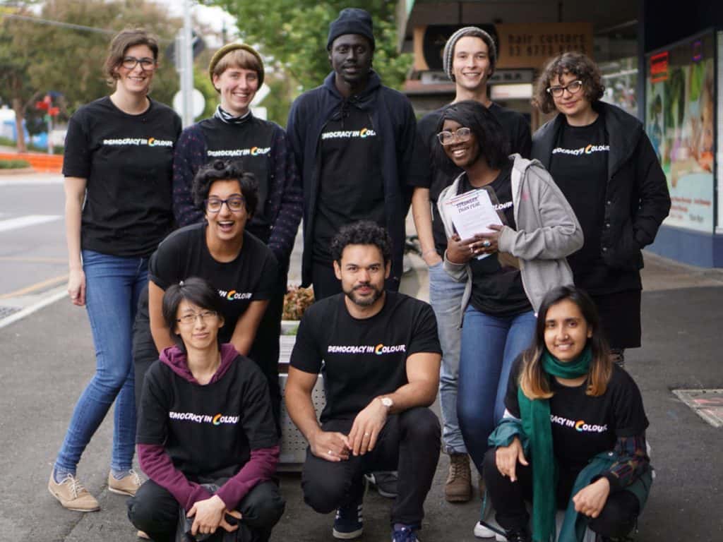 A group of people of different races stand together. Each person wears a Democracy in Colour tshirt.