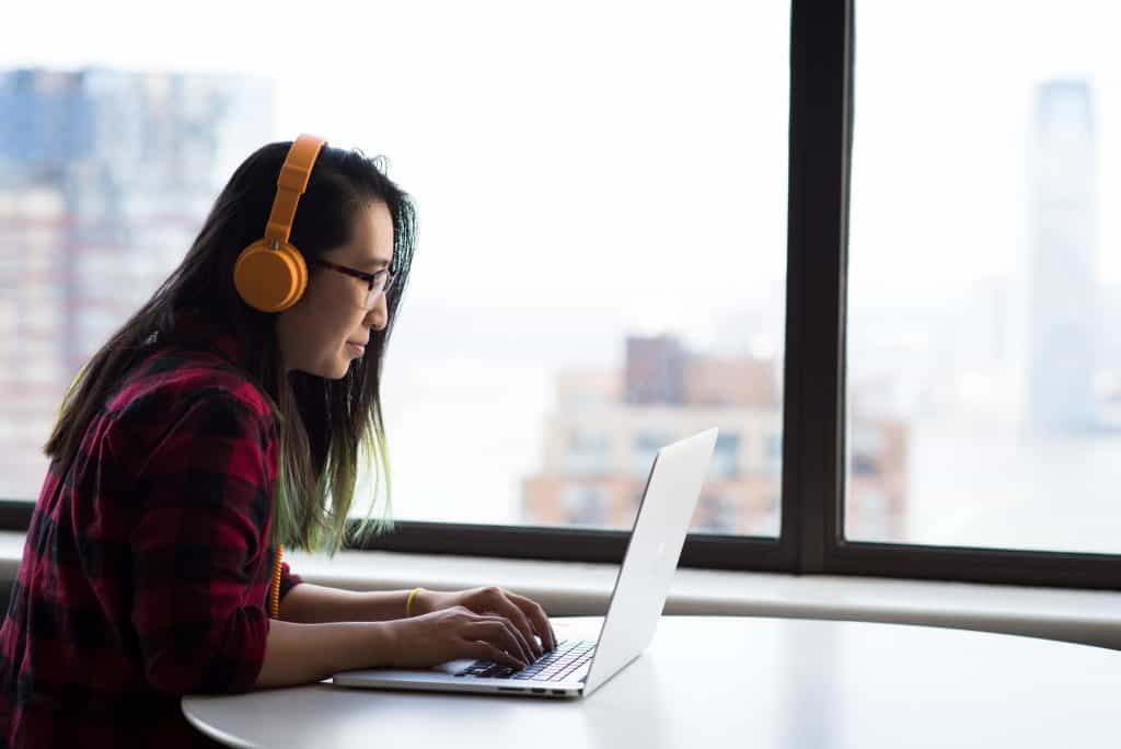 A woman sits at a laptop, wearing headphones. She sits in front of a window showing a city skyline.