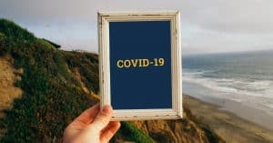 Photograph of a hand holding a wooden picture frame, with a cliff, beach and sea showing in the backgroun. 'COVID-19' is written inside the frame.