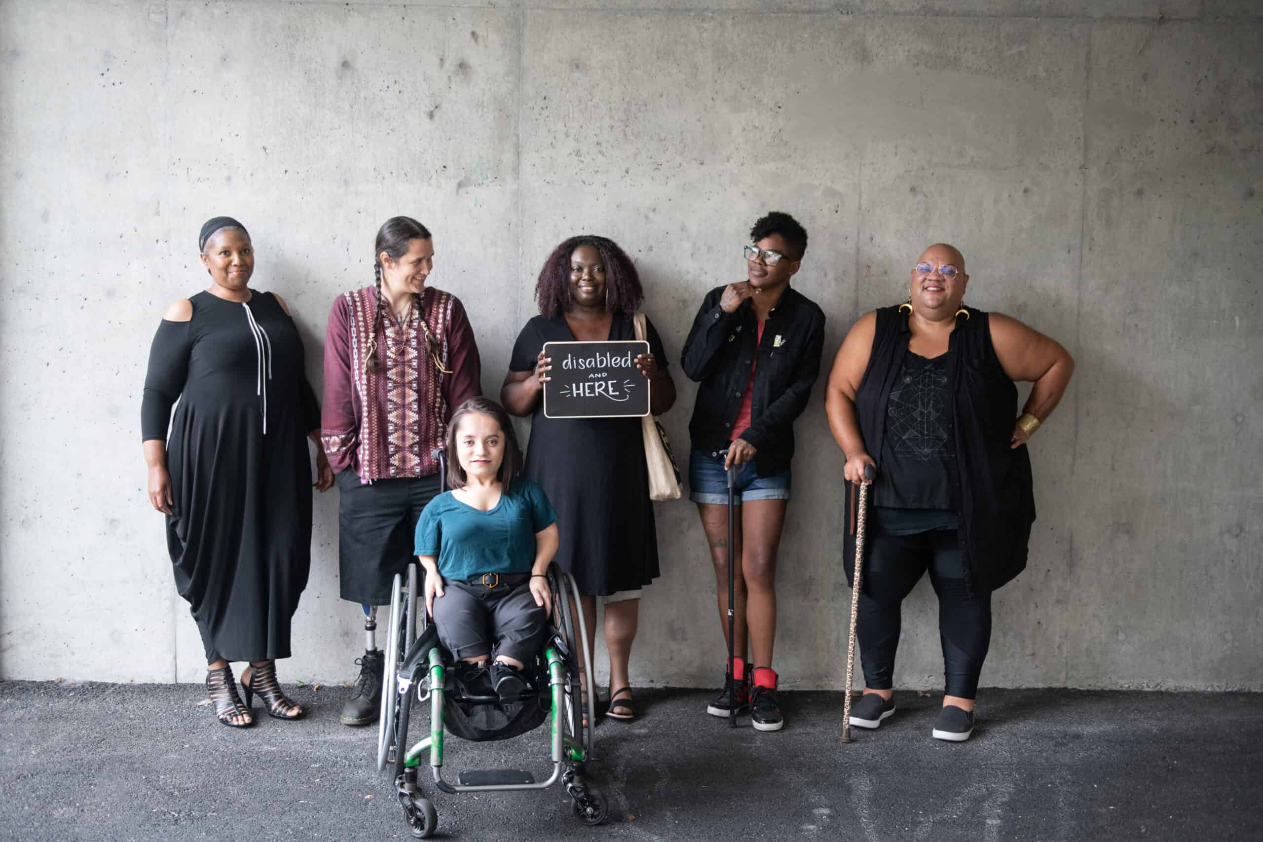 Six disabled people of color smile and pose in front of a concrete wall. Five people stand in the back