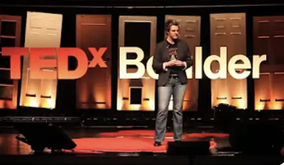 Erica Chenoweth giving a TED talk on stage. Behind Erica is a sign that reads TEDx Boulder'.