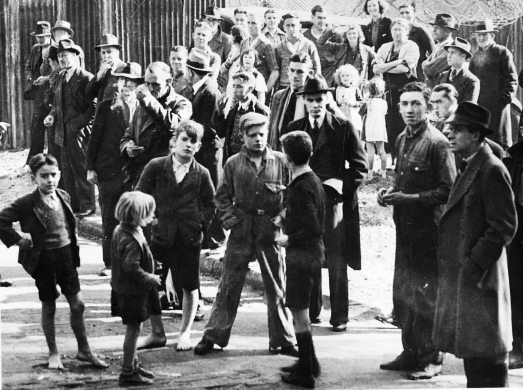 Black and white photo of a crowd of people including several children.|Photograph of a billboard advertising Winfield cigarettes. The billboard advertising reads 'Anyhow... Winfield 25s'. Graffiti has been inserted so it reads 'Anyhow buga up a Winfield 25'.