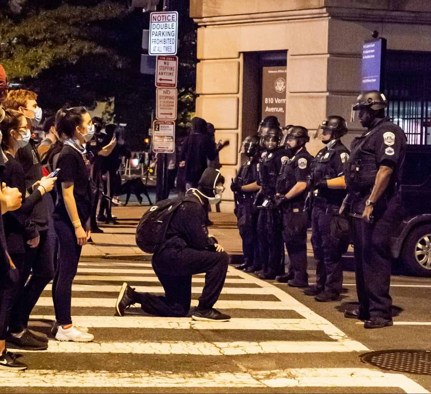 Protestors in a line with one protestor kneeling on knee (in honour of George Floyd) in front of line of police at protest in street in Washington