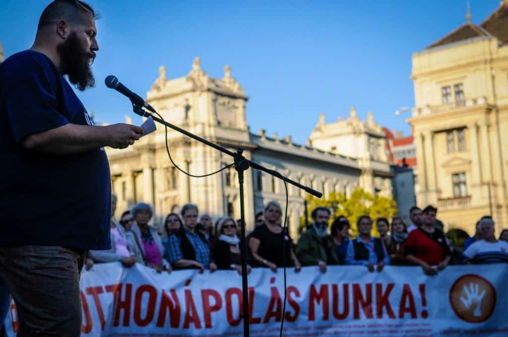 Máté speaks into a microphone at a protest outside the Hungarian Parliament. In the background protestors hold a large poster with Hungarian text.