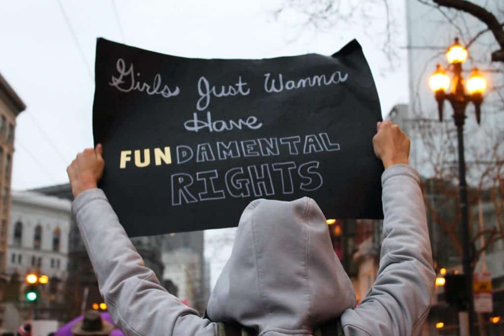 Photo of a woman at a rally taken from behind. She is wearing a grey hoodie sweatshirt and holding a sign that reads 'Girls just want to have FUNdamental rights'