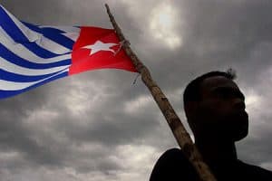 The Morning star flag, symbol of West Papuan independence, is flying against a grey cloudy sky. The person who is holding it is in silhouette.