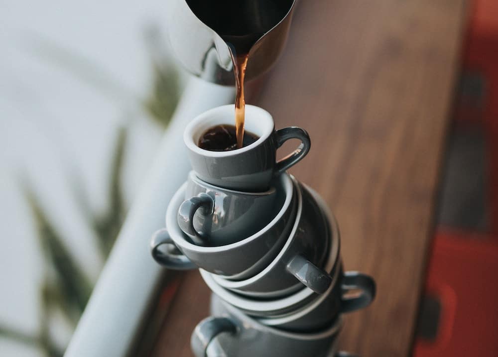 Coffee is poured from a metal jug into a large and unstable stack of coffee cups sitting on a wooden table.
