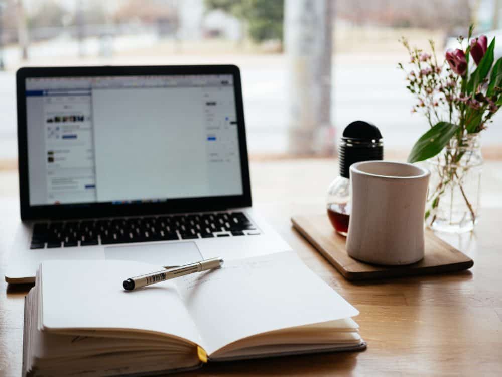 A desk in a brightly lit room, with a laptop, coffee and coffee cup, and a notebook and pen sitting on it.
