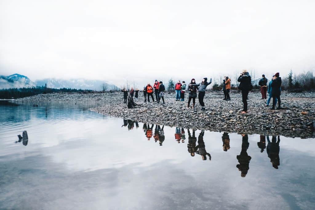 A small group of people stand on the beach of a beautiful lake, wearing winter clothing