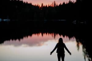 The silhouette of a person standing by a lake at sunset. The trees on the other side of the lake are reflected in the water, and the overall effect is calm but a bit lonely.