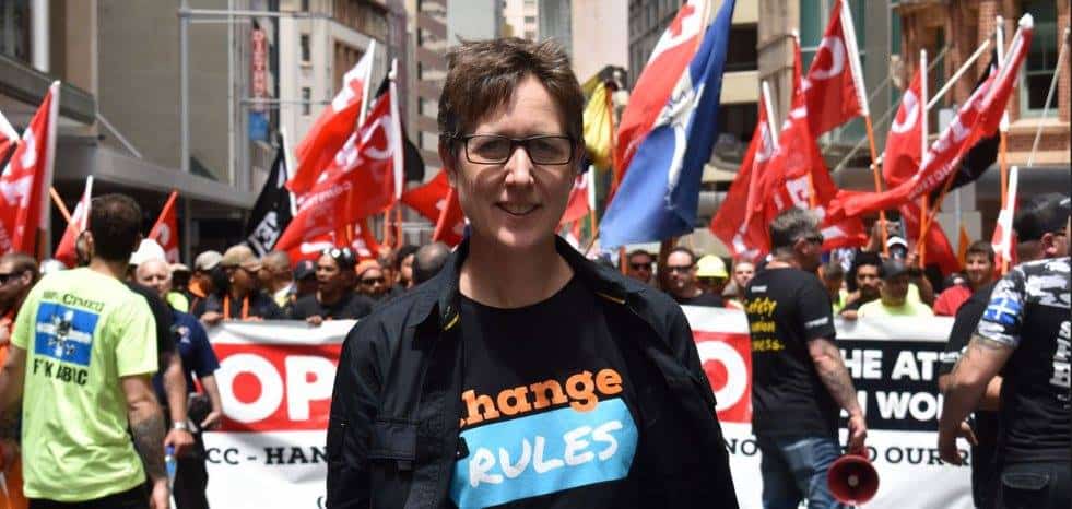 Sally McManus wears a 'Change the Rules' tshirt in front of a large crowd of workers holding banners and flags.