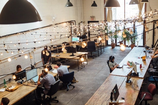 People sitting in a large room on shared desks in front of computer monitors with fairy lights above.