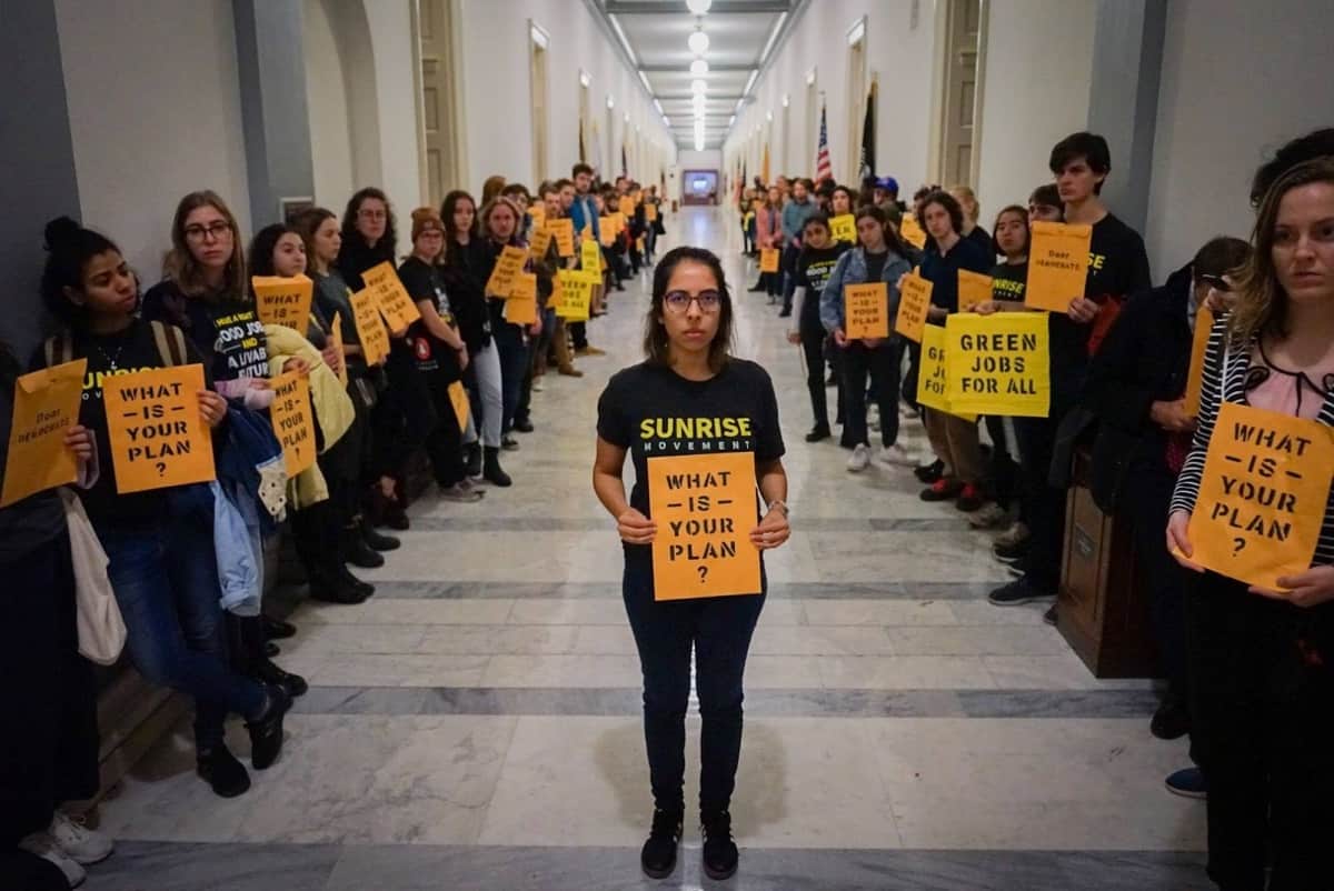young people from the Sunrise Movement lining the walls of a large walkway inside a building. They are holding signs that say What is your plan? There is one woman standing at the front holding an orange piece of paper that reads What is your plan?
