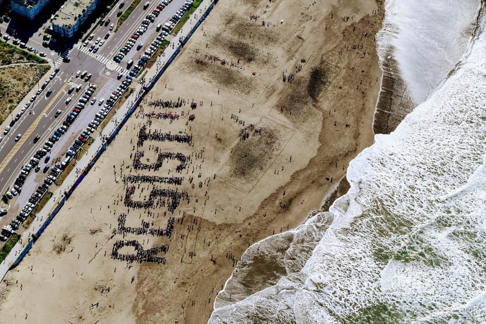 A birds-eye view of Ocean Beach in California, with thousands of people gathered above the waves to spell out RESIST!! There are two exclamation marks, one of which is mostly pink, indicating it is made up of feminist activists.