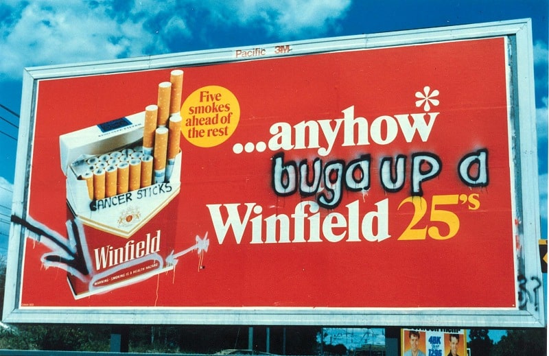 Photograph of a billboard advertising Winfield cigarettes. The billboard advertising reads 'Anyhow... Winfield 25s'. Graffiti has been inserted so it reads 'Anyhow buga up a Winfield 25'.