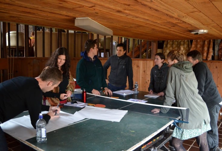 A group of people stand together around a ping pong table covered in paper and pens.